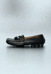 Mocassin en cuir noir avec un détail pompon, des coutures visibles et des picots en caoutchouc sur la semelle, présenté en vue latérale sur une surface grise.