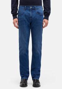 Niebieskie dżinsy z prostą nogawką, z pięcioma kieszeniami, metalowym guzikiem i subtelnym przetarciem dodającym faktury. Widoczne czarne buty.