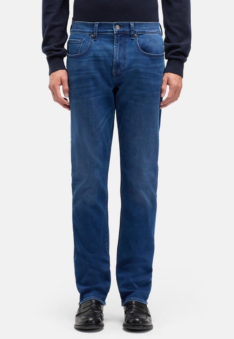 Niebieskie dżinsy z prostą nogawką, z pięcioma kieszeniami, metalowym guzikiem i subtelnym przetarciem dodającym faktury. Widoczne czarne buty.