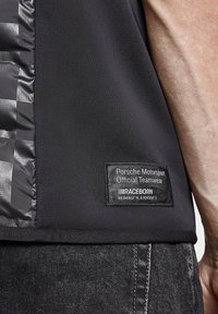 Czarny sportowy top z teksturowanym bocznym panelem, z metką "Porsche Motorsport Official Teamwear" oraz wyszytymi współrzędnymi.