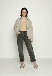 Casaco de ganga oversized de areia claro, top de alças castanho, jeans verdes escuros com bainha e sandálias beijas com ferragens decorativas. Conjunto simples e descontraído.