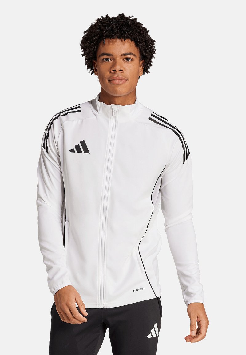 Jeune homme portant une veste de sport blanche zippée avec des bandes noires et un pantalon de sport noir avec un logo blanc, debout sur un fond uni.