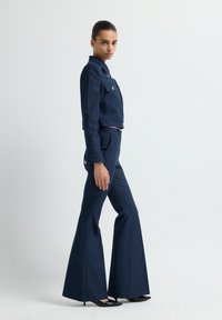 Námořnická modrá croptop bunda se dvěma předními kapsami a zlatými knoflíky, zkombinována s kalhotami s vysokým pasem a zvonovými nohavicemi. Hladká textura, přiléhavý střih.