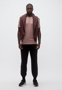 Άνδρας όρθιος, φοράει πουκάμισο μοβ, καφέ φούτερ πάνω στους ώμους, μαύρο φόρμες και μαύρα αθλητικά παπούτσια σε λευκό φόντο.