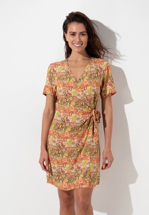 Donna sorridente con lunghi capelli scuri indossa un vestito corto a maniche corte con motivi paisley arancioni, verdi e rosa, in piedi contro un muro bianco.