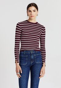 Frau, die ein maroon-rot und weiß gestreiftes Langarmoberteil und hochgeschossene blaue Jeans mit vorderen aufgesetzten Taschen trägt.