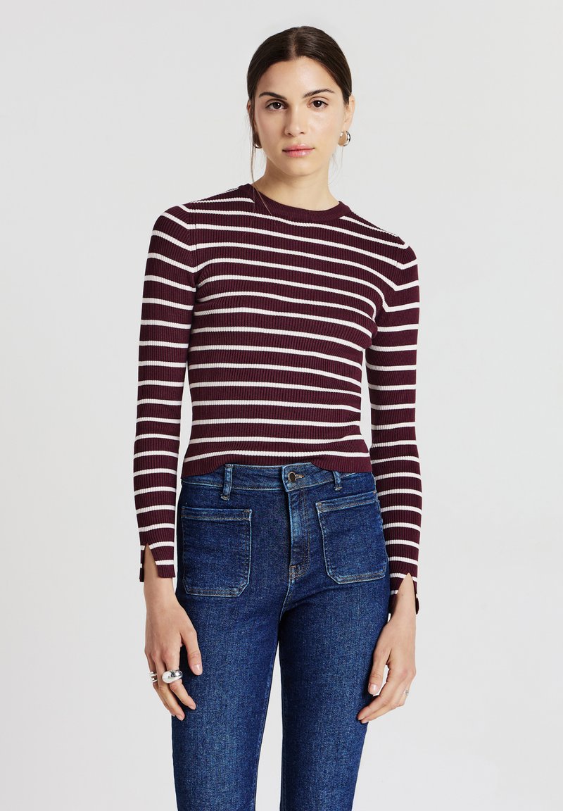 Frau, die ein maroon-rot und weiß gestreiftes Langarmoberteil und hochgeschossene blaue Jeans mit vorderen aufgesetzten Taschen trägt.