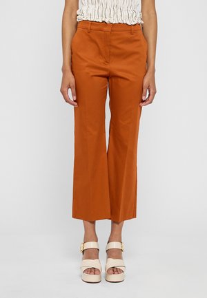 Femme portant un pantalon taille haute, coupe large, couleur orange brûlé et des sandales à plateforme crème avec des boucles dorées sur un fond clair.