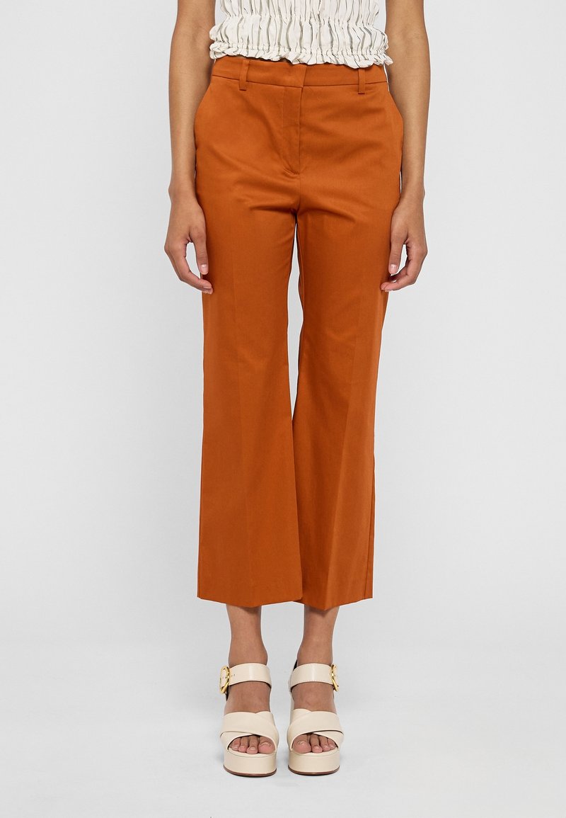 Femme portant un pantalon taille haute, coupe large, couleur orange brûlé et des sandales à plateforme crème avec des boucles dorées sur un fond clair.