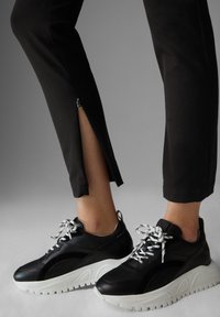 Benen in zwarte broek met zijritsen en zwarte chunky sneakers met witte zolen en veters op een grijze achtergrond.