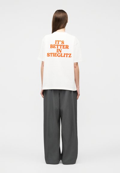 Personne aux longs cheveux bruns portant un T-shirt blanc avec un texte orange "C'EST MIEUX À STIEGLITZ" et un pantalon gris ample, debout de dos.