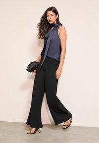 Sötétkék, ujjatlan felső, redőzött nyakkivágással és hosszú megkötővel. Magas derekú fekete, bő szárú nadrág. A modell egy kis fekete clutch-ot tart.