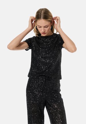 Jonge vrouw die haar haar rechtzet, draagt een zwart topje met korte mouwen en pailletten en bijpassende broek met grote zwarte oorringen, effen achtergrond.