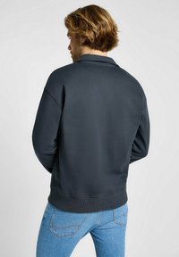Felpa grigia scura con una vestibilità rilassata, polsini e orlo a coste, e una texture liscia. Indossata con jeans azzurri chiari, che mostrano la vista posteriore.
