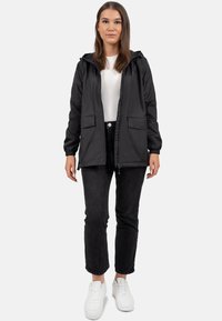 Giubbotto nero con cappuccio, polsini elastici, due tasche frontali, chiusura con zip, indossato su una maglietta bianca e jeans neri. Sneakers bianche.