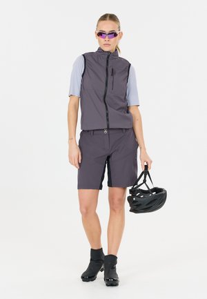 Vrouw draagt een grijze fietsvest en korte broek, paarse zonnebril, zwarte laarzen, houdt een zwarte fietshelm naast zich vast.