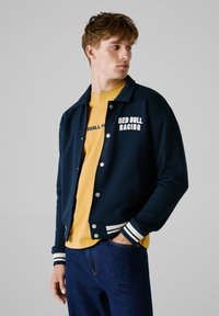 Marine sweatshirtjakke med hvide stribede manchetter, trykknapper og teksten "RED BULL RACING". Bæres over en gul t-shirt, parret med blå jeans.