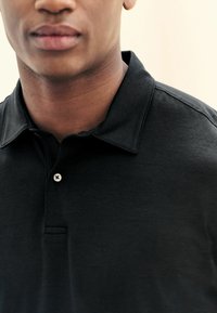Camisa polo negra con cuello, que presenta un solo botón blanco y una tela suave y texturizada, con enfoque en el área del cuello y la parte superior del pecho.