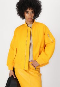 Giacca bomber gialla brillante con chiusura a zip, polsini a coste e tasche laterali; presenta una tasca con zip sulla manica e una texture liscia.