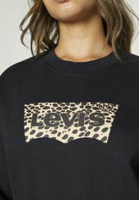 Crni dukserica sa uzorkom leopard u boji logoa Levi's na prednjoj strani. Ima okrugli izrez i rebraste manžete, izrađena je od mekog materijala.