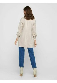 Cappotto beige a maniche lunghe in materiale morbido, con una vestibilità rilassata e spalle scese, abbinato a pantaloni in denim blu larghi e stivaletti crema.
