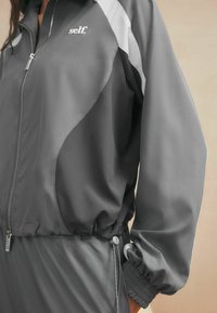 Gray zip-up jacket made of smooth material with white accents. Features elastic cuffs and a drawstring at the waist. Logo on the chest.