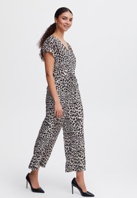 Combinaison à motifs léopard avec des manches courtes, taille cintrée et pantalon à jambes larges, associée à des talons aiguilles noirs. Fond clair.