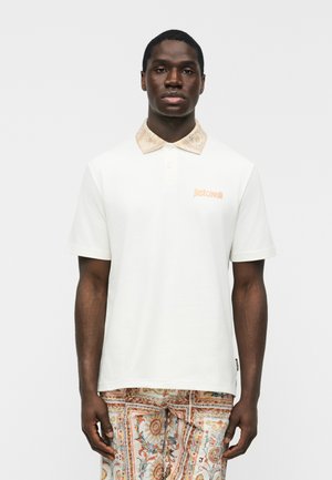 Bărbat purtând un tricou polo alb cu mâneci scurte, cu un guler cu model și pantaloni cu același model, pe un fundal simplu, deschis la culoare.