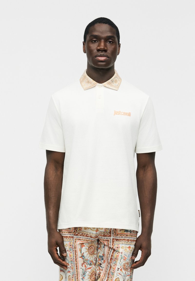 Homme portant un polo blanc à manches courtes avec un col à motifs et un pantalon assorti à motifs, sur un fond uni clair.