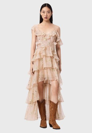 Kobieta w różowej, warstwowej, przejrzystej sukience w kwiaty z asymetrycznym dołem i falbanami, nosząca brązowe zamszowe kowbojskie buty, stoi na tle jednolitego tła.