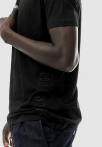 T-shirt nera a maniche corte con una texture morbida e un logo in rilievo verticale lungo il lato, abbinata a jeans scuri.