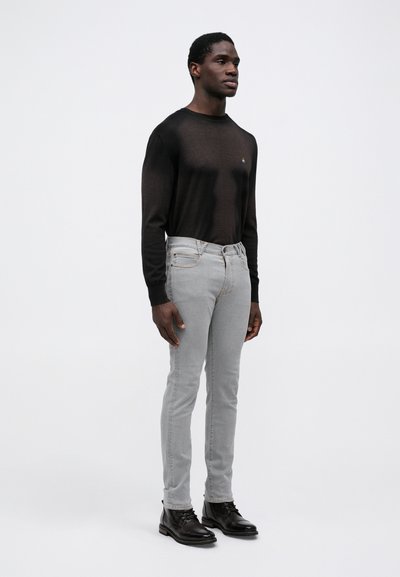 Črna dolga majica z dolgimi rokavi, ožji svetlo sivi jeans in črne čipkaste škornje. Diskreten logo na majici, gladka tekstura, minimalističen dizajn.