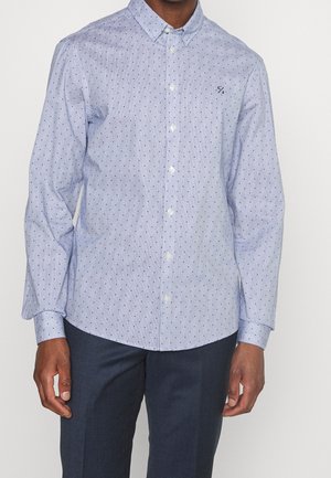 Man draagt een lichtblauw overhemd met kleine donkere polkadots en een donkere broek, staand tegen een effen witte achtergrond.