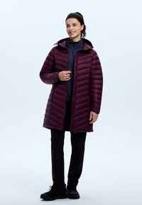 Bordeaux pufferjack met een capuchon, verticale gestikte patroon, rits aan de voorkant en zakken, gedragen over een blauwe top en zwarte broek.