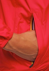 Rode nylon jas met elastische manchetten, met een zijzak met een kleurrijke gestreepte ritsdetail en een hand gestoken in de zak.