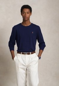 Νεαρός άνδρας φορώντας ναυτικό μπλε πουκάμισο μακρύ μανίκι με μικρό λογότυπο, λευκά παντελόνια ψηλής μέσης, καφέ ζώνη και ρολόι χειρός, με τα χέρια στις τσέπες.