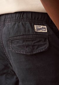 Donkerblauwe broek met een gestructureerde afwerking, met een achterzak en een geweven label waarop "Superdry Athletic Goods" staat. Elastische tailleband.