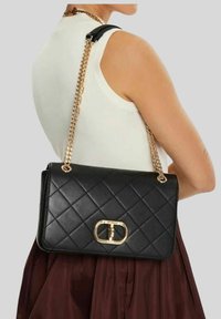 Sac à main noir matelassé avec un fermoir en métal doré, équipé d'une bandoulière en chaîne et d'accents en cuir, reposant contre une jupe bordeaux.