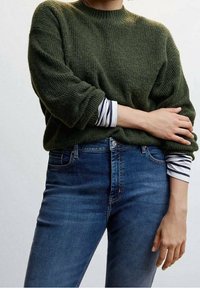 Person wearing a green knit sweater over a white and navy striped long-sleeve shirt, paired with blue denim jeans, arms crossed.