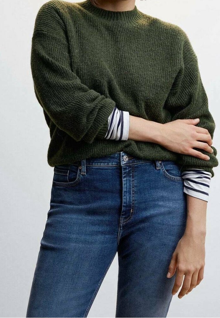 Person wearing a green knit sweater over a white and navy striped long-sleeve shirt, paired with blue denim jeans, arms crossed.