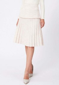 Femme portant une jupe midi plissée blanche, un haut à manches longues blanc et des escarpins blancs à bout pointu, se tenant devant un fond uni.