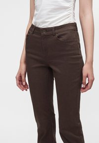 Femme portant un pantalon marron foncé ajusté et un haut blanc sans manches froncé, debout devant un fond clair uni.