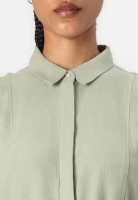 Light green collared shirt made from textured fabric, featuring a single pearl button, with vertical seams for a structured look.