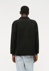 Jachetă verde închis din catifea cu guler clasic și mâneci lungi, văzută din spate, asortată cu blugi din denim albastru deschis.