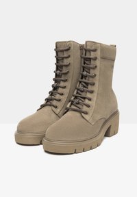 Beige ruskindsnits snørebånd støvler til ankler med en chunky hæl, der har metaløjer, teksturerede snørebånd og en sidelukning for nem påklædning.