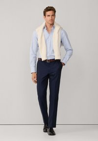 Tenue masculine comprenant une chemise rayée bleu clair et blanche, un pull crème drapé sur les épaules, un pantalon bleu marine et des chaussures noires.