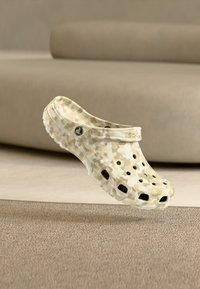Μπεζ και λευκά Crocs με μοτίβο καμουφλάζ που επιπλέουν εσωτερικά πάνω από μοκέτα με μπεζ καναπέ στο φόντο.
