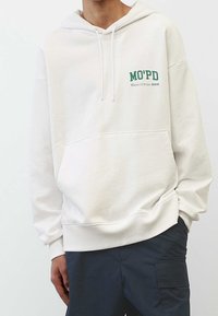 Sudadera beige claro hecha de tela suave, con un gran bolsillo frontal y texto del logo en verde. Corte oversize y detalle de capucha con cordón.