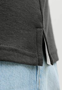 Gros plan sur l'ourlet d'une chemise noire avec une fente latérale, porté sur un jean en denim bleu clair, montrant la texture du tissu et les coutures détaillées.