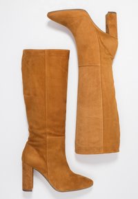 Par de botas altas de ante color beige con tacones gruesos, una bota de pie y la otra acostada sobre un fondo blanco.